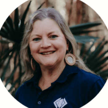 A woman smiling outdoors wearing a navy shirt and circular woven earrings, surrounded by trees